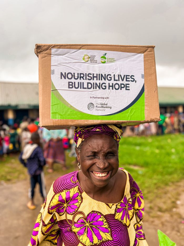 Elderly woman joyfully holding a donation box on her head outdoors, spreading hope.